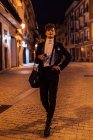 Jovem transgênero na moda masculino com a mão na cintura passeando no pavimento urbano e olhando para a câmera no crepúsculo — Fotografia de Stock