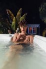 Junger transsexueller Mann mit langen Nägeln blickt in der Abenddämmerung in die Kamera, die im Whirlpool liegt — Stockfoto