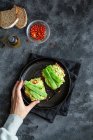 Top view crop anonymous female holding tasty fresh toasts with guacamole and green peas served on black plate — Stock Photo