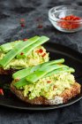 Torradas apetitosas com guacamole fresco e vagens de ervilhas verdes decoradas com pimentas vermelhas e servidas em prato preto — Fotografia de Stock