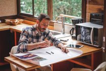 Серьезный мужчина-архитектор, сидящий за столом на кухне и рисующий чертеж здания карандашом и линейкой, работая дома — стоковое фото