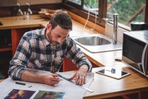 Серйозний чоловічий архітектор сидить за столом на кухні і малює план будівлі з олівцем і лінійкою під час роботи вдома — стокове фото