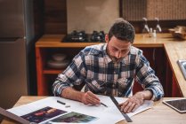 Arquitecto masculino serio sentado a la mesa en la cocina y dibujando el plano del edificio con lápiz y regla mientras trabaja en casa - foto de stock