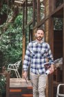 Серьезный бородатый мужчина-архитектор с рулоном бумаги и планшетом, стоящий возле современного деревянного дома в лесу и смотрящий в камеру — стоковое фото