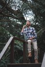 Seitenansicht eines seriösen männlichen Architekten, der auf einem Geländer auf einer Holzterrasse sitzt und während der Diskussion über das Projekt mit dem Smartphone spricht — Stockfoto