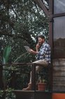 Vista lateral del arquitecto masculino serio sentado en la barandilla en la terraza de madera y hablando en el teléfono inteligente mientras discute el proyecto - foto de stock