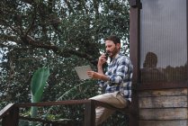 Vue latérale d'un architecte masculin sérieux assis sur une balustrade sur une terrasse en bois et parlant sur un smartphone tout en discutant du projet — Photo de stock
