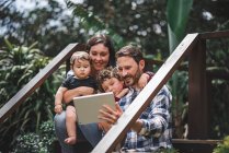 Coppia allegra con bambini carini seduti su gradini di legno di casa e scattare selfie su tablet — Foto stock