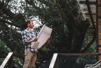 Vista laterale a basso angolo di architetto maschio con progetto di carta in piedi sulla terrazza della casa in legno nella foresta — Foto stock