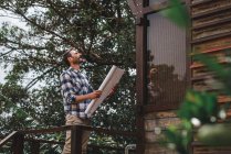 Низкий угол зрения архитектора мужского пола с бумажным черновиком, стоящим на террасе деревянного дома в лесу — стоковое фото
