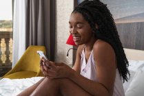Щаслива молода афроамериканка з довгим кучерявим волоссям у сонному одязі посміхається і читає повідомлення на смартфоні, відпочиваючи на зручному ліжку. — стокове фото