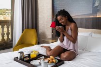 Corpo pieno di contenuti giovane afroamericana turista donna in camicia da notte seduta su un comodo letto con vassoio di deliziosa colazione e bere caffè in hotel moderno — Foto stock
