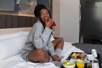 Полное содержание молодая афроамериканская туристка в ночной одежде, сидящая на удобной кровати с трапезой вкусного завтрака в современном отеле, глядя в сторону — стоковое фото
