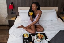 Dall'alto corpo pieno di contenuti giovane afroamericana turista donna in camicia da notte seduta su un comodo letto con vassoio di deliziosa colazione e bere caffè in hotel moderno guardando altrove — Foto stock