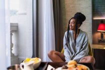 Volle Länge der positiven jungen ethnischen Reisenden sitzt auf einem Stuhl im Hotelzimmer und trinkt während des Frühstücks an sonnigen Tagen frischen Saft — Stockfoto