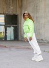 Vista lateral de cuerpo completo de la joven afroamericana con el pelo rizado teñido con elegante chaqueta verde con pantalones blancos y botas de moda mirando hacia otro lado mientras está de pie en la calle urbana pavimentada - foto de stock