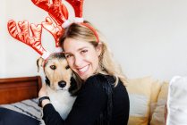 Счастливая женщина в декоративных оленьих рогах обнимает очаровательную чистокровную собаку, глядя в камеру на кровати — стоковое фото