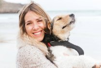 Cheerful woman hugging cute purebred dog while looking at each other against sea under cloudy sky — Stock Photo