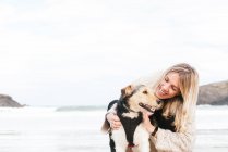 Seitenansicht der Frau umarmt niedlichen reinrassigen Hund, während sie einander gegen das Meer unter bewölktem Himmel — Stockfoto