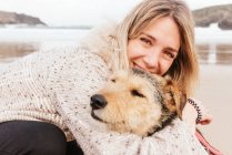 Cheerful woman hugging cute purebred dog while looking at each other against sea under cloudy sky — Stock Photo