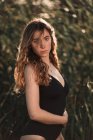 Side view of charming young brunette with long wavy hair wearing black swimsuit looking at camera while standing against blurred green trees in sunny garden — Stock Photo