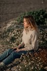 Side view of calm contemplative young female traveler in casual wear sitting alone on flowering meadow against blurred plowed field while relaxing in countryside in spring day — Stock Photo