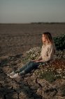 Visão lateral do corpo inteiro da calma contemplativa jovem viajante em desgaste casual sentado sozinho no prado florido contra o campo arado borrado enquanto relaxa no campo no dia da primavera — Fotografia de Stock