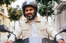 Von unten der brutale ethnische männliche Biker mit Helm, der zeitgenössisches Motorrad fährt, während er sich auf die Stadt freut — Stockfoto