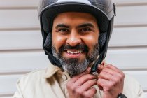 Fröhlicher männlicher Motorradfahrer mittleren Alters setzt Schutzhelm auf und blickt in Kamera gegen gerippte Wand — Stockfoto