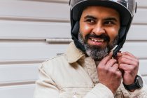 Веселий середнього віку етнічний чоловічий мотоцикліст одягає захисний шолом, дивлячись на камеру проти ребристої стіни — стокове фото