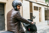 Вид збоку жорстокого етнічного байкера чоловічої статі в шоломі, що керує сучасним мотоциклом, дивлячись в місто — стокове фото