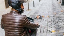 Back view of brutal ethnic male biker in helmet driving contemporary motorbike while looking way in town — Stock Photo