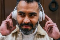 Кроп дружественных бородатый этнический мужчина слушать песню из беспроводной гарнитуры, глядя в сторону при дневном свете — стоковое фото