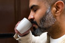 Вид сбоку на культуру концентрированного среднего возраста этнического мужчины с закрытыми глазами, наслаждающегося ароматом горячего напитка в чашке — стоковое фото