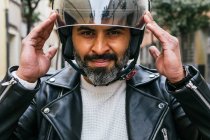 Веселий середнього віку етнічний чоловічий мотоцикліст одягає захисний шолом, дивлячись на камеру проти ребристої стіни — стокове фото