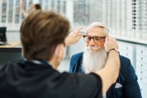 Back view of professional optician in mask helping senior male putting glasses while working in optical store — Stock Photo