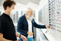 Side view of optician in mask and senior male choosing eyeglasses and sunglasses in optical store — Stock Photo