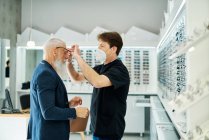 Side view of professional optician in mask helping senior male putting glasses while working in optical store — Stock Photo