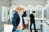 Вид збоку професійного оптика в масці допомагає старшому чоловікові вибирати окуляри під час роботи в оптичному магазині — стокове фото