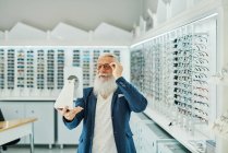 Contenido elegante hombre mayor mirando en el espejo mientras se prueba y la elección de gafas en la tienda óptica moderna - foto de stock