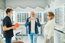 Optician with mirror helping content elderly male trying on eyeglasses in optical store — Stock Photo