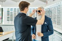 Side view of professional optician in mask helping senior male putting sunglasses while working in optical store — Stock Photo