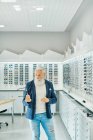 Serious elderly bearded male in trendy outfit standing with modern stylish glasses in optical shop — Stock Photo