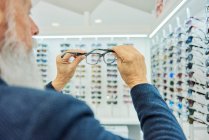 Серьезный пожилой бородатый мужчина в модном наряде, стоящий с современными стильными очками в оптическом магазине — стоковое фото