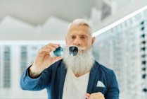 Serious elderly bearded male in trendy outfit standing with modern stylish sunglasses in optical shop — Stock Photo