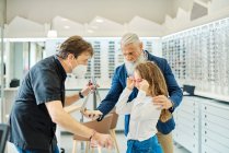 Side view of teenage girl trying on glasses while standing in optical store with grandfather and optician and looking in mirror — Stock Photo