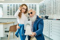 Веселий старший чоловік тримає дзеркало в той час як дівчина-підліток намагається на окулярах в сучасному оптичному магазині — стокове фото