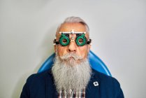 Бородатий літній чоловік в окулярах для тестування очей сидить у сучасній клініці перед оглядом зору і дивлячись на камеру — стокове фото