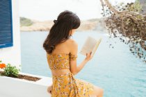 Вид збоку на романтичну молоду жінку в літньому вбранні сидить на терасі біля озерної води і читає цікавий роман у книзі, проводячи літній час у сільській місцевості — стокове фото