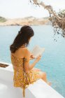 Вид сбоку романтичной молодой женщины в летнем наряде, сидящей на террасе у воды озера и читающей интересный роман в книге, проводя лето за городом — стоковое фото